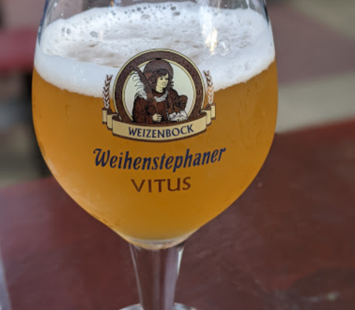 A glass of Weihenstephaner Vitus beer, golden in color, with a logo featuring a character on the front.