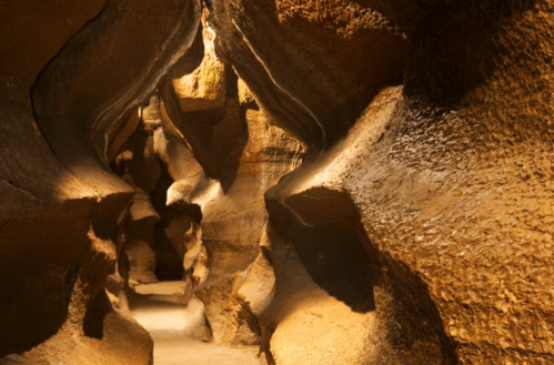 A narrow, winding cave passage with textured, illuminated rock walls and a sandy floor.