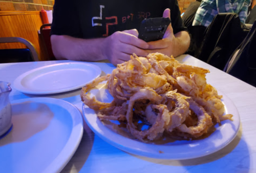 A plate of crispy onion rings sits on a table next to a person using a smartphone.