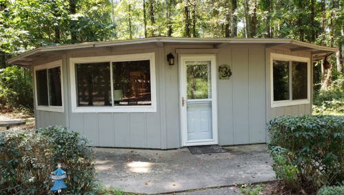 A small, gray, circular cabin surrounded by trees, featuring large windows and a front door.