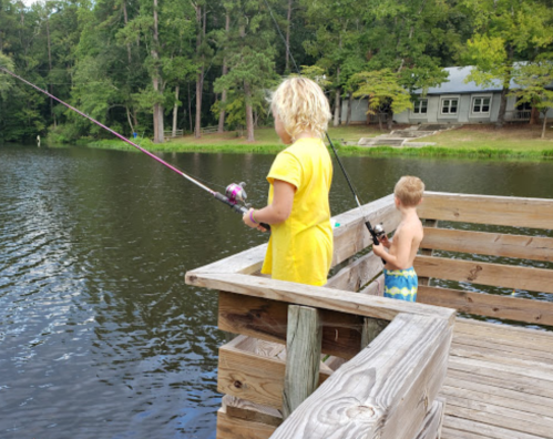 Two children fishing from a wooden dock by a lake, surrounded by trees and a house in the background.