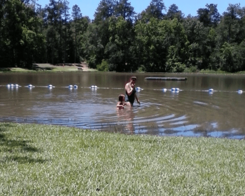 Two people wade in a calm lake surrounded by trees, with a grassy area in the foreground.