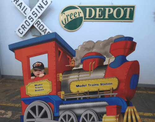 A child peeks out from a colorful train cutout at a model train station display.