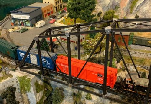 A model train crosses a black metal bridge over a scenic landscape with buildings and cars in the background.