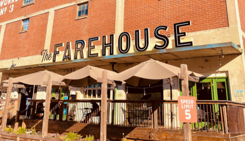 Exterior of The Farehouse restaurant with a wooden deck, umbrellas, and a speed limit sign. Bright, sunny day.