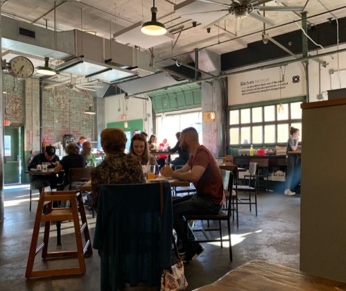 A bustling café interior with patrons seated at tables, exposed brick walls, and large windows letting in natural light.