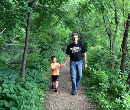A man and a young boy walk hand in hand along a lush, green path surrounded by trees.