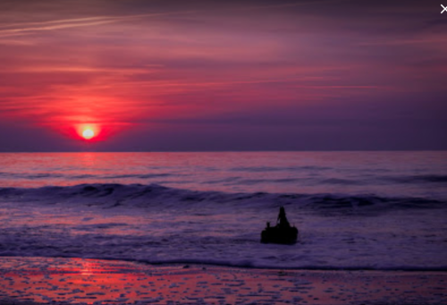 A serene sunset over the ocean, with gentle waves and a silhouette of a person sitting on the shore.