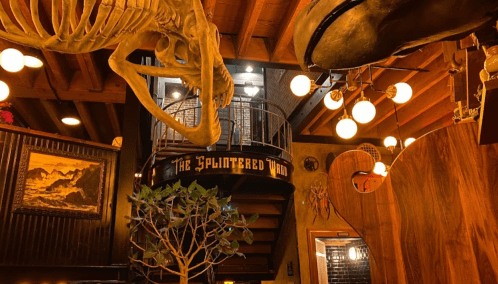 Interior of a rustic restaurant featuring a dinosaur skeleton, warm lighting, and a spiral staircase.
