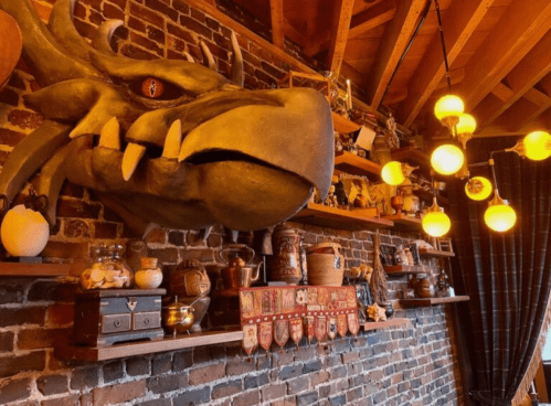A rustic interior featuring a dragon head mount, shelves with various collectibles, and warm pendant lights.