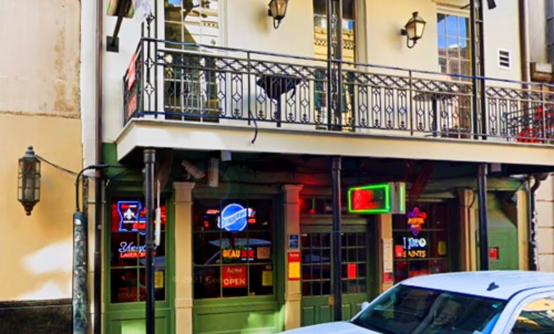 A colorful bar exterior with neon signs, a balcony, and a parked car in front.