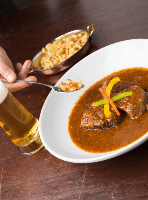 A plate of beef in sauce garnished with peppers, alongside a bowl of pasta and a glass of beer.