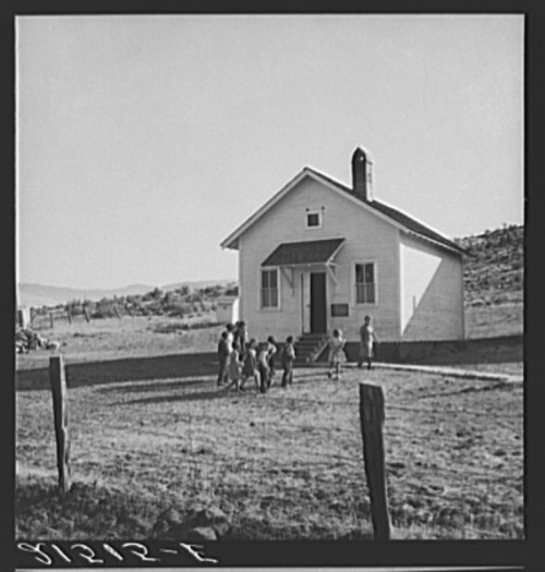 A small white schoolhouse with children playing outside on a grassy field, surrounded by a rural landscape.