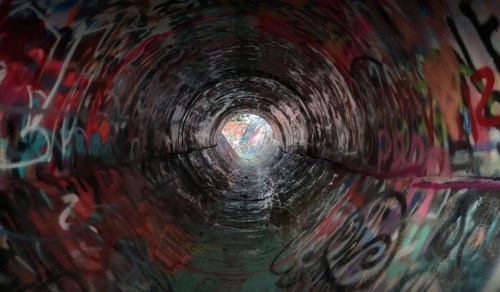 A view inside a graffiti-covered tunnel, leading to a bright opening at the far end.