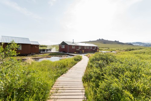 A wooden pathway leads to a red cabin surrounded by lush greenery and mountains under a bright sky.