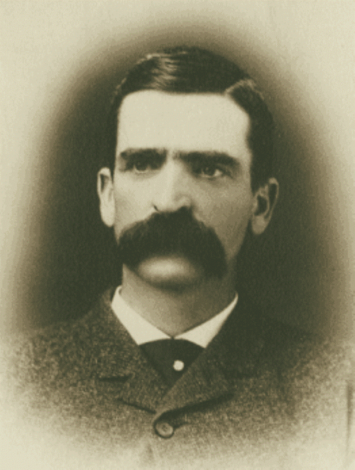 Black and white portrait of a man with a prominent mustache, wearing a suit and looking directly at the camera.