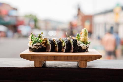 A wooden platter holds a colorful sushi roll, garnished with sprouts, against a blurred outdoor background.