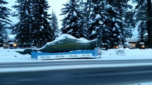 A large, green dinosaur statue covered in snow, with trees and a building in the background.