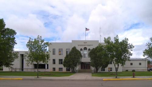 Sheridan County building with a flag, surrounded by trees and a grassy area under a cloudy sky.