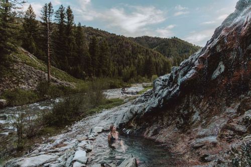 A person relaxes in a natural hot spring surrounded by rocky terrain and lush green mountains under a blue sky.