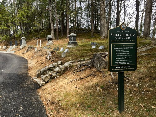 Sign welcoming visitors to Sleepy Hollow Cemetery, surrounded by gravestones and trees along a winding path.