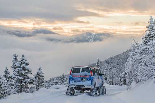 A vehicle on a snowy mountain road, surrounded by trees and clouds, with a sunset in the background.