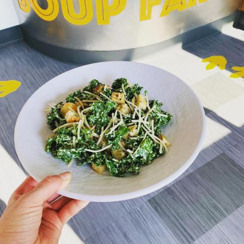 A hand holding a bowl of kale salad with croutons and cheese, set against a colorful background.