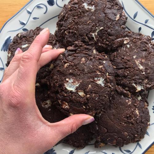 A hand reaching for a plate of chocolate cookies with white chocolate chunks, arranged on a decorative dish.