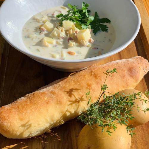 A bowl of creamy soup with potatoes and herbs, accompanied by a crusty bread roll and fresh potatoes on a wooden board.