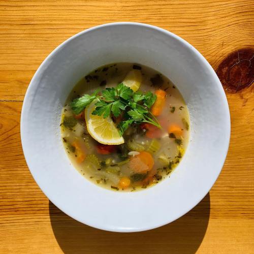 A bowl of vegetable soup with carrots, celery, and herbs, garnished with a lemon slice and fresh parsley.