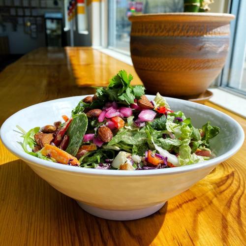 A vibrant salad in a white bowl, featuring greens, nuts, and colorful toppings, set on a wooden table by a window.