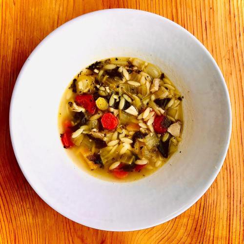 A bowl of soup with orzo, vegetables, and pieces of chicken, served on a wooden table.