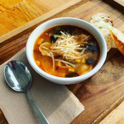 A bowl of soup topped with cheese, served with a slice of bread on a wooden tray. A spoon rests beside the bowl.