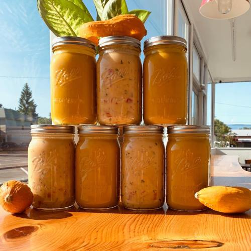 A stack of seven jars filled with colorful preserves, with a squash and green leaves in the background.