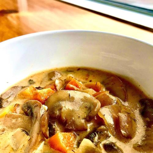 A close-up of a creamy mushroom soup with carrots and greens in a white bowl, set against a wooden background.