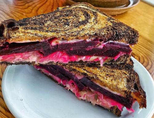 A close-up of a sliced sandwich with dark rye bread, filled with layers of beets and sauerkraut on a white plate.