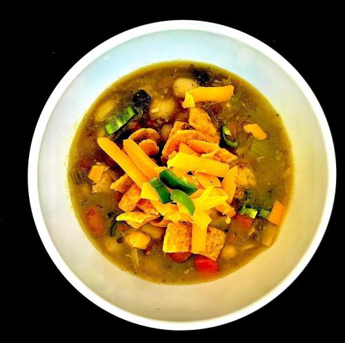 A bowl of green chili soup topped with cheese, tortilla strips, and colorful vegetables on a black background.