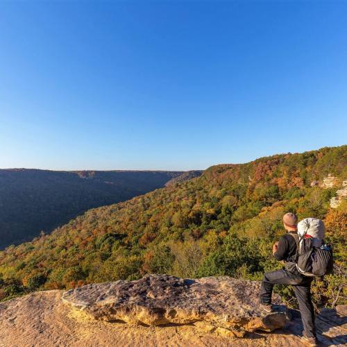 A hiker stands on a rocky ledge, overlooking a colorful valley with trees in autumn hues under a clear blue sky.