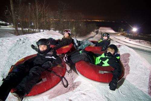 Four kids in winter gear relax on snow tubes at night, with a snowy slope and city lights in the background.