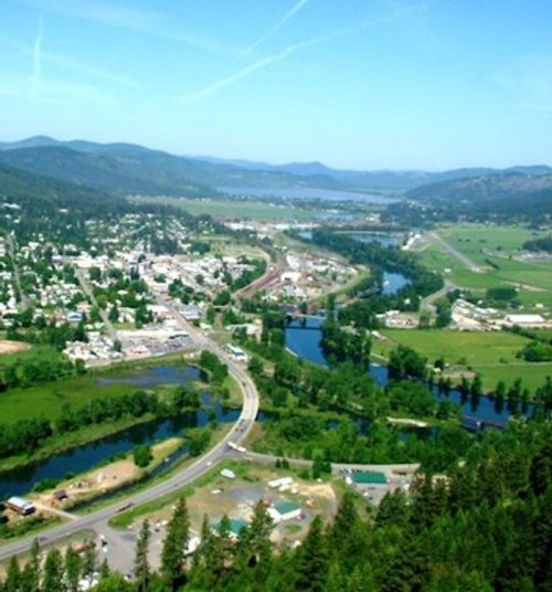 Aerial view of a lush green valley with a river, roads, and a small town surrounded by mountains under a clear blue sky.