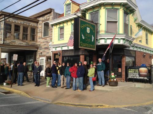A group of people stands outside a colorful Irish pub, waiting in line on a sunny day.