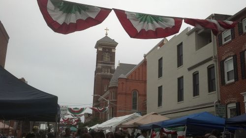 A street festival scene with colorful bunting, tents, and a church tower in the background under a cloudy sky.