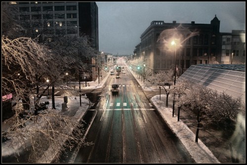 A snowy city street at dusk, with traffic lights glowing and vehicles navigating through the winter scene.