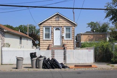 A small, shingled house with a white door, surrounded by a fence and trash bags on the sidewalk. Clear blue sky above.