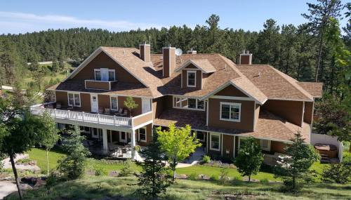 A large, multi-story house surrounded by trees and greenery, featuring a balcony and a well-maintained lawn.