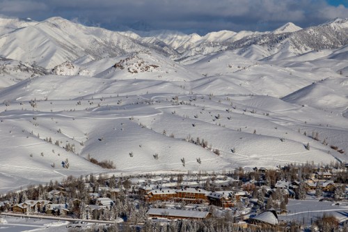 Snow-covered mountains and hills surround a small town nestled in a valley, under a partly cloudy sky.
