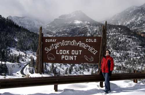 A person in a red jacket stands beside a wooden sign reading "Switzerland of America Look Out Point" with snowy mountains in the background.