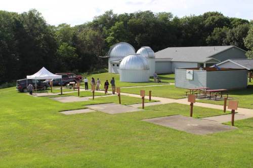 A grassy area with observatory domes, a building, and people at shooting stations under a clear sky.