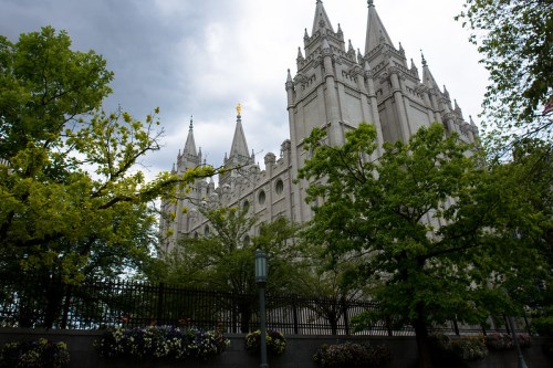 A tall, ornate stone building with spires, surrounded by green trees and flowers under a cloudy sky.