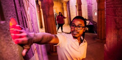 A person with glasses reaches out to touch a colorful wall in a dimly lit, ancient-themed environment.